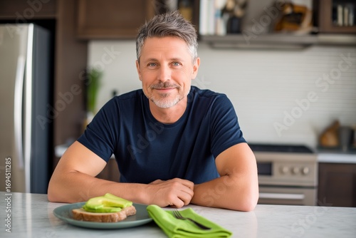 Smiling middle-aged man sitting at kitchen counter with healthy avocado toast. Mature male enjoying fresh breakfast in bright modern home interior.
