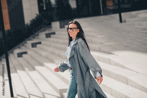 Mature Woman in Grey Trench Coat Walking Outdoors in Modern Urban Setting on a Sunny Day