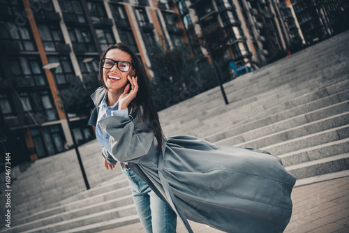 Attractive young brunette woman with glasses, wearing a stylish trench coat, enjoying an urban outdoors leisure day