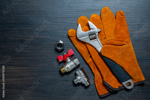 Adjustable wrench, ball valve with red handle, stainless steel tee fittings, leather safety gloves and rubber gaskets on wooden background, top view, copy space, closeup. Plumbing repair concept