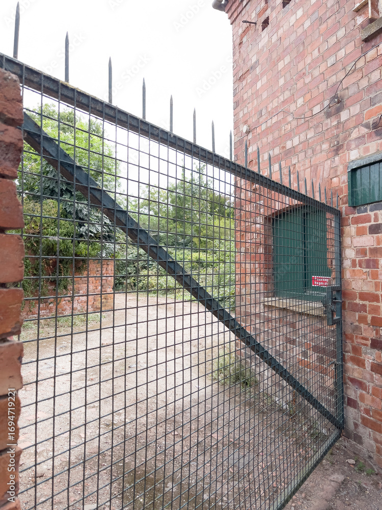 Fototapeta premium A large, industrial metal gate against a red brick building, offering a view into an overgrown, outdoor area