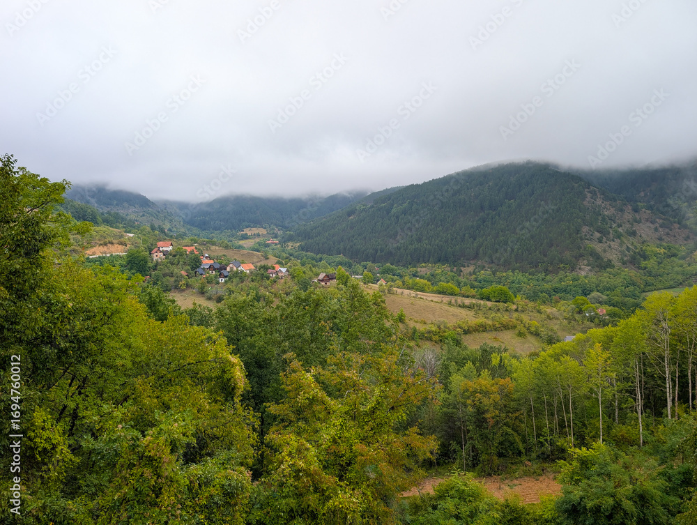 Fototapeta premium Foggy mountains and a valley with small houses and red roofs among green trees. Peaceful rural living and natural beauty in the Balkans.