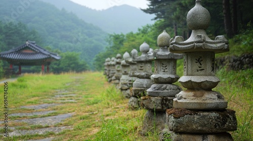 Wallpaper Mural A row of stone lanterns with intricate designs, standing in a grassy field with a traditional Korean pavilion in the background, under a cloudy sky with mountains Torontodigital.ca