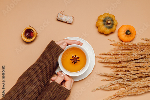 Herbal tea with spices and anise in woman hands with autumn decorations, pumpkin and pampas grass top view. Hands hold tea on brown background