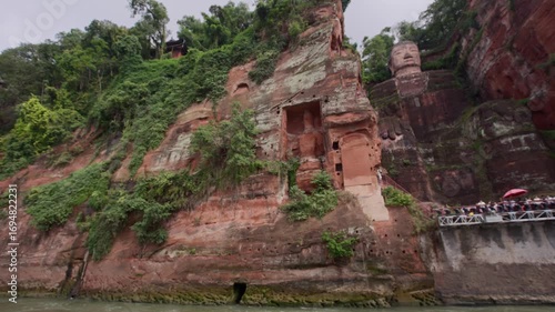 Wide view of Leshan Giant Buddha and cliff base with walking tourists near riverbank in slow motion
