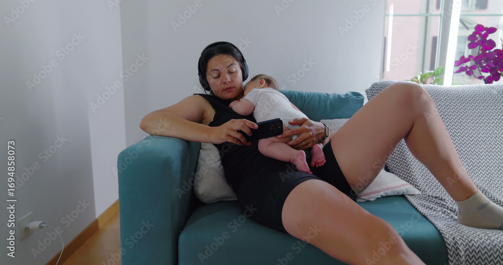 Obraz premium Mother and baby relaxing on the couch, the baby lies comfortably on her chest while she uses her phone, capturing a serene and intimate family moment in a cozy home environment-SD 480p