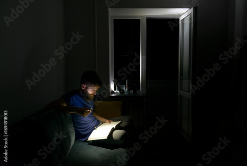 Man reading book with flashlight during power outage at night, sitting on sofa in dark room