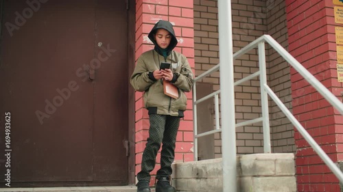 Wallpaper Mural Little boy in hooded jacket stands by building column holding brown book in one hand while operating phone with focus, urban background with red and white brick wall and staircase railing Torontodigital.ca