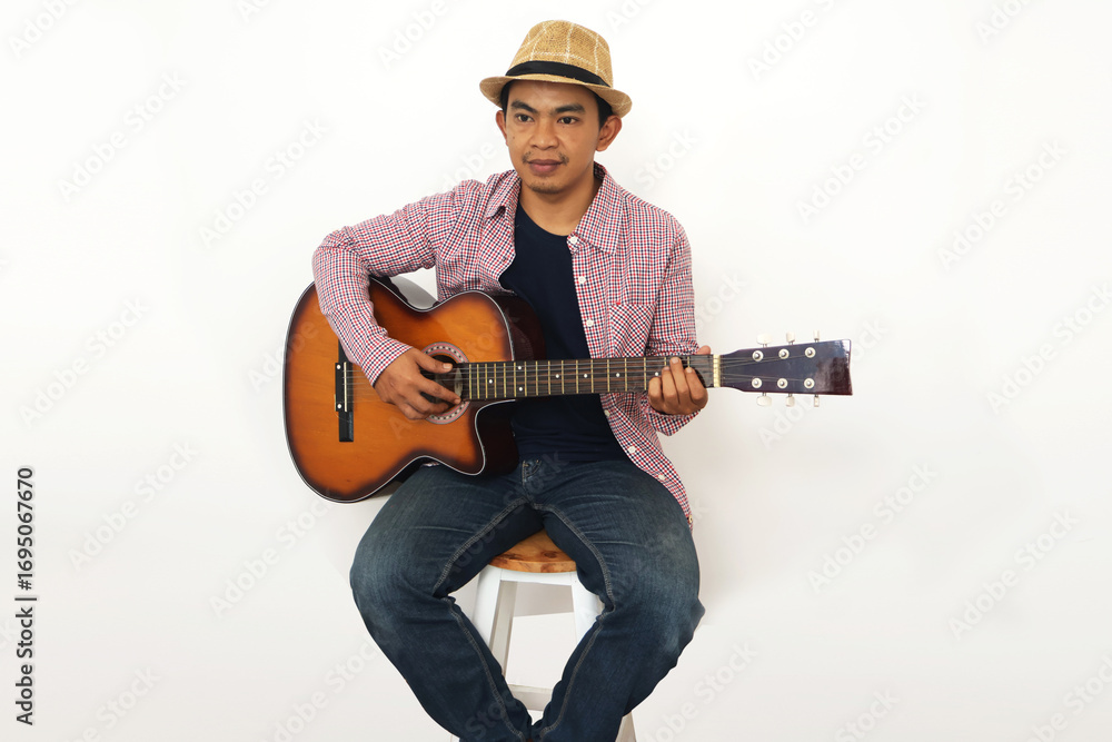 Obraz premium Young Asian man in brown cowboy hat and checkered shirt holding acoustic guitar against white background