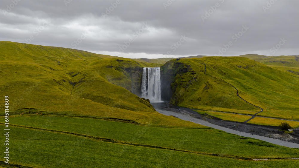 Fototapeta premium Skogafoss waterfall flows powerfully from a cliff, surrounded by green hills and a misty spray. A winding path and dark riverbed complete the dramatic scene.