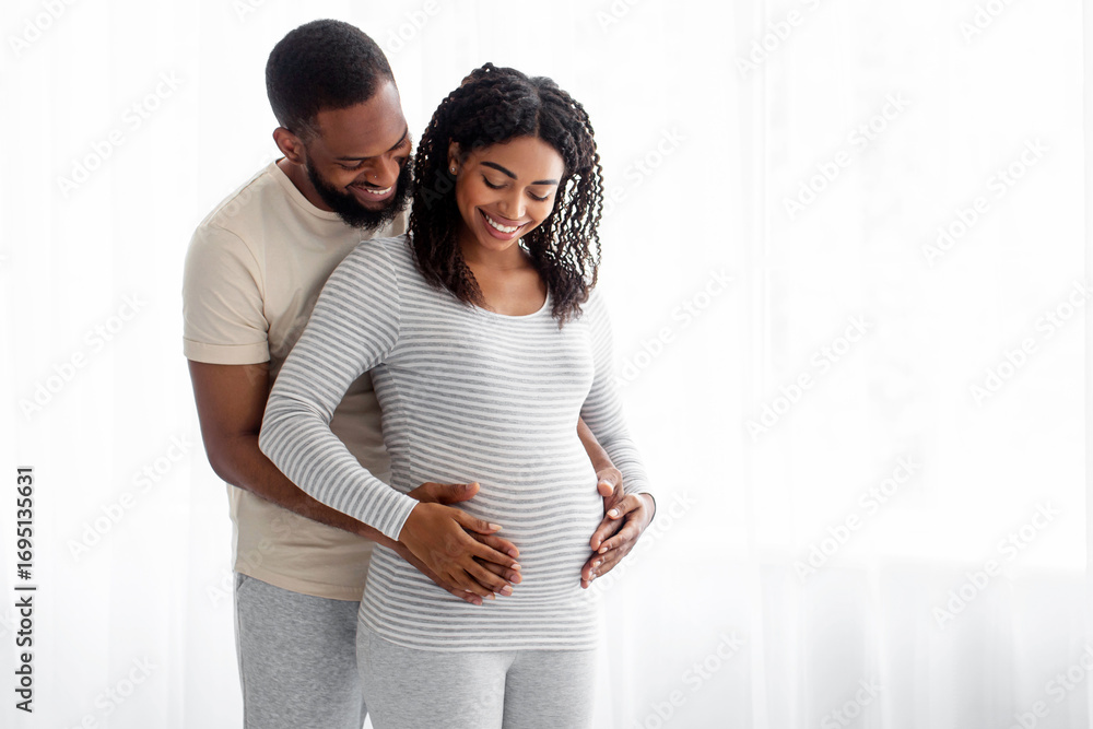 Fototapeta premium Satisfied millennial black handsome hugs pregnant wife in bedroom interior and look at belly on window background. Couple awaiting baby, cute parents-to-be together at home, family enjoy tender moment