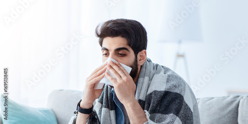Cuadro en lienzo Ill arab man covered in warm blanket looking at laptop screen and sneezing nose in paper tissue, sitting on sofa at home, empty space