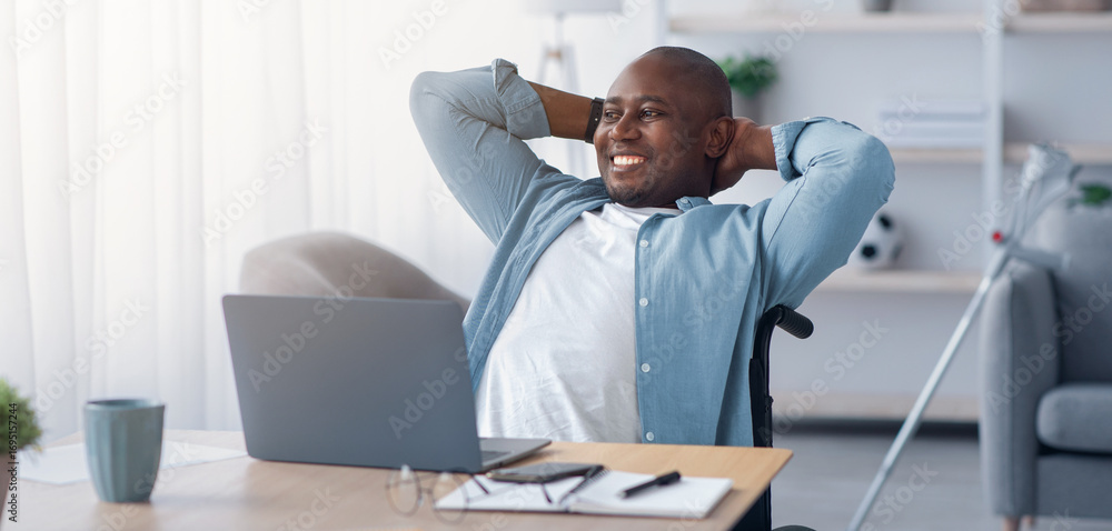 Obraz premium Rest and relax at home, work remotely, successful business and break. Happy mature african american guy disabled in wheelchair threw hands behind head at table with laptop in living room interior