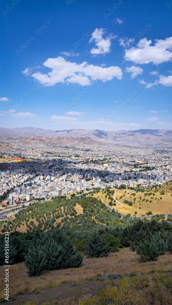 Obraz premium Panoramic view of Sanandaj city from Abidar heights, showcasing urban sprawl amid mountain ranges and greenery under a partly cloudy sky. 
