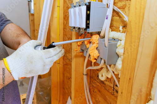 An electrician installs wiring outlets while sealing gaps with spray foam insulation in new home being built.
