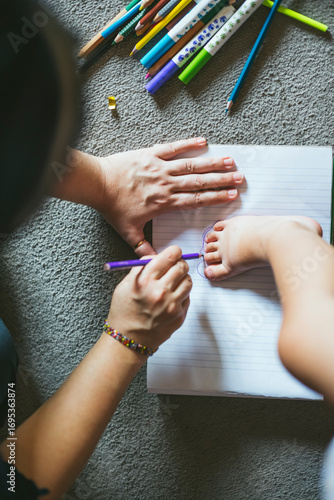 Parent tracing baby’s foot on notebook with colored pencils