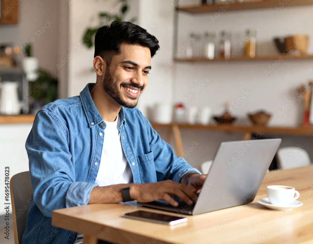 Fototapeta premium Cheerful young adult man in casual outfit at cafe, laptop open and coffee on table, smiling as he works, creative freelancer or startup scene, stylish business lifestyle stock photography.