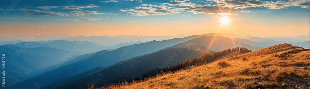 Obraz premium Scenic mountain range view at sunrise. Golden grass slope in foreground, blue hazy mountains, cloudscape, & bright sun