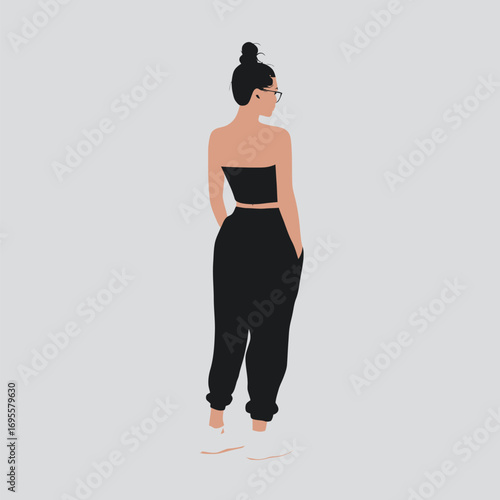 A confident woman stands with her back to the viewer, showcasing a trendy black outfit that highlights her stylish appearance. She sports a bun hairstyle and glasses.