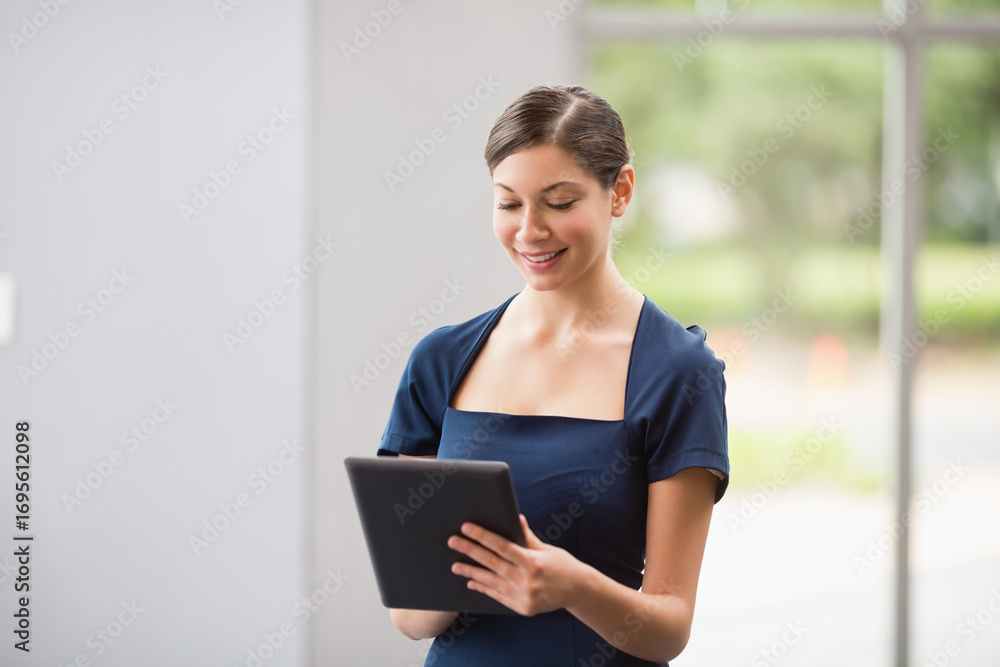 Obraz premium Woman standing in office lobby wearing navy blue dress holding black tablet and scrolling by window