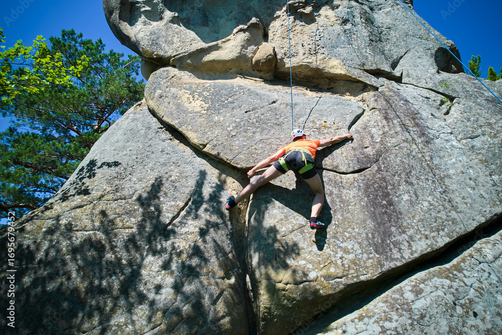 Obraz premium Aerial view of male rock climber ascending rugged limestone cliff with harness and rope for safety. Sportsman climbing on vertical large boulder at Dobvush Rocks in Carpathian mountains, Ukraine.