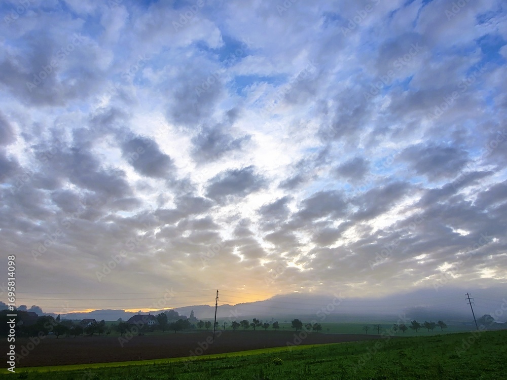 Obraz premium Scenic morning atmosphere with the sun rising over the countryside, dramatic cloud formations in the sky, lush green meadows, and gentle morning mist on the horizon