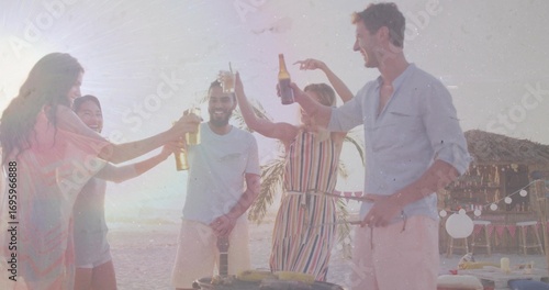 Friends raising beer bottles and toasting barbecue on sunset beach, with grill and string lights