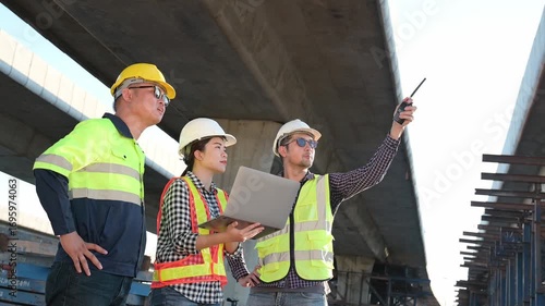A team of Asian engineers in safety helmets and vests using laptop and walkie-talkie at construction site under bridge, symbolizing teamwork, communication, and project management.