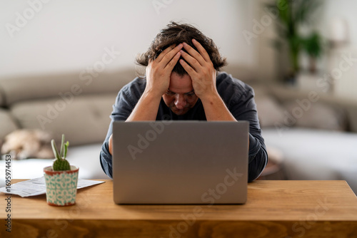 A stressed person with hands on head sits at a wooden desk with a laptop. A cactus plant and papers add to the modern home office setting, conveying frustration and work pressure.