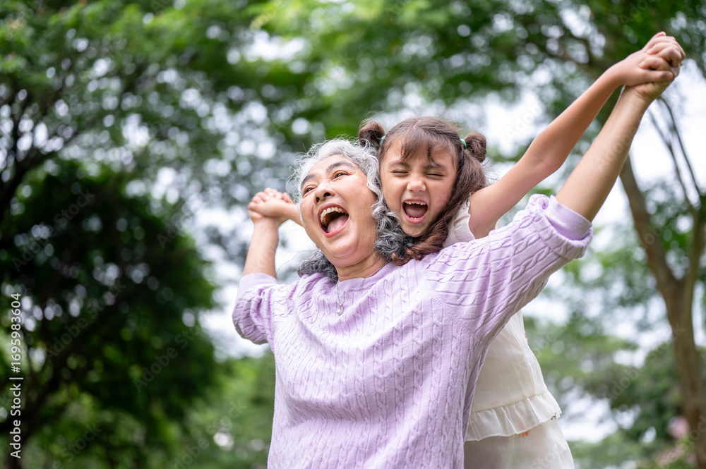 Naklejka premium Laughing old woman grandmother playing with a kid girl granddaughter behind her back in the park.