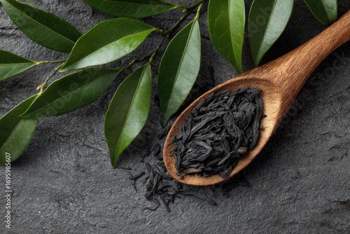 Wooden spoon holding loose black tea leaves, surrounded by fresh green leaves on a dark gray stone surface
