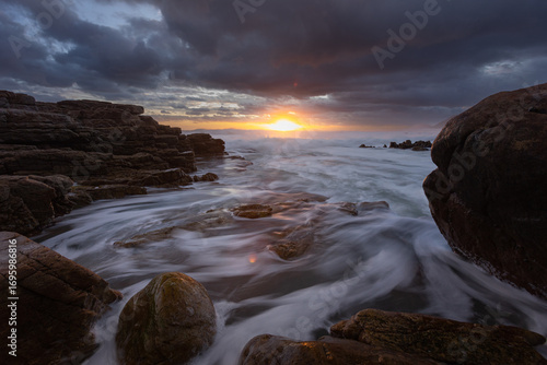 View of turbulent waters swirling around rugged rocks under a dramatic sky ablaze with the setting sun's golden light, Cape Town, Western Cape, South Africa.