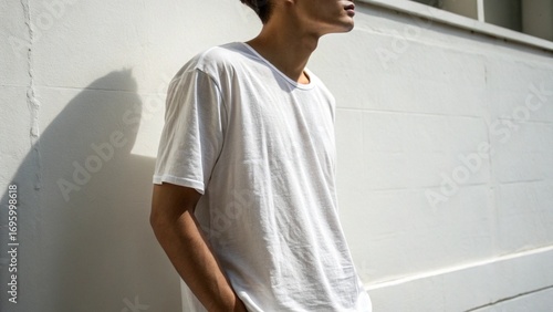 Young man wearing plain white t shirt mockup leaning on wall outdoors in sunlight 
