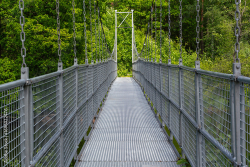 A long and narrow suspension bridge, supported by strong chains, winds its way through vibrant green foliage, Sweden, Knared