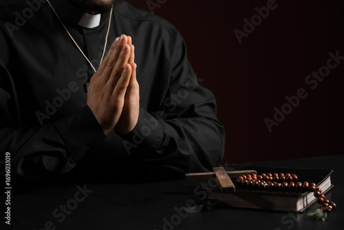 Young priest with wooden cross, beads and Bible praying at table on red background, closeup