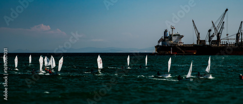 A scene from a sailboat race at sea. Numerous small Optimist-class sailboats are visible, each with 