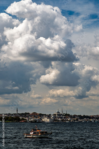A small boat sails alone on the sea, with Istanbul's historic mosques and the city skyline in the background. Heavy clouds and dramatic light in the sky.