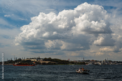 A view of the Bosphorus. Topkapi Palace and Hagia Sophia are visible on the left, and the Blue Mosque is on the right. A small boat is in the foreground, and a cloudy sky is in the background.