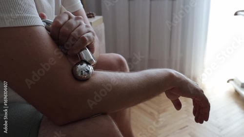 A close-up of a man's arm being massaged with a metallic roller, highlighting self-care, pain relief, and wellness. The scene is set indoors with natural light