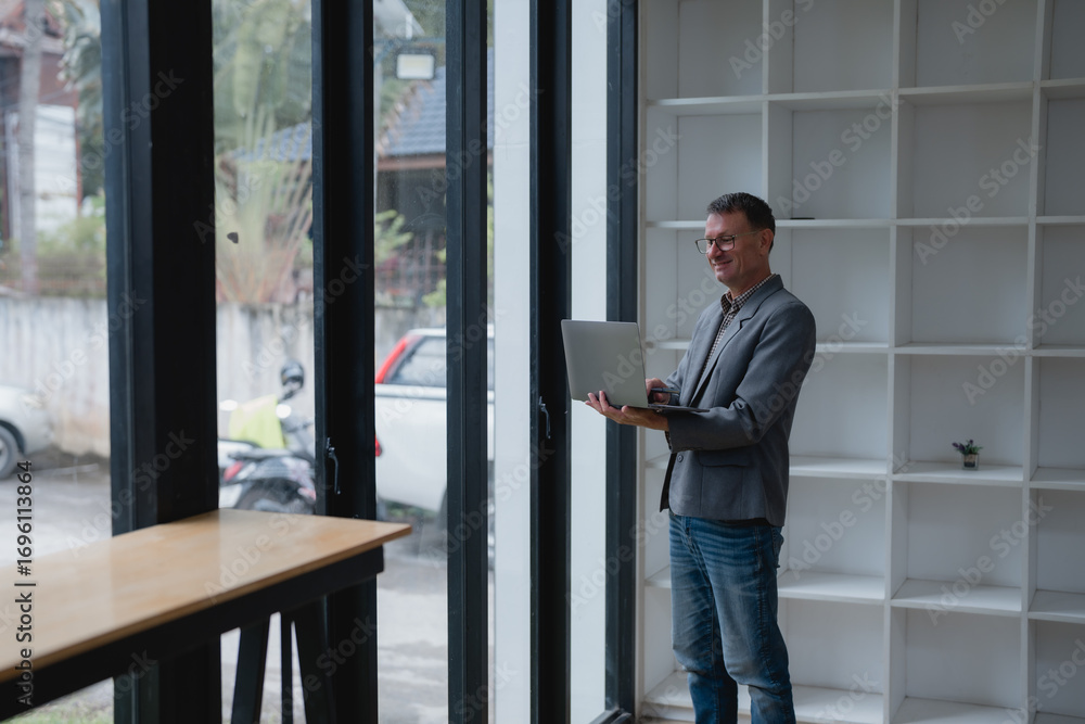 Fototapeta premium Businessman wearing glasses and a grey jacket, standing and working on his laptop in a modern office space filled with natural light, smiling happily while engaging in his tasks