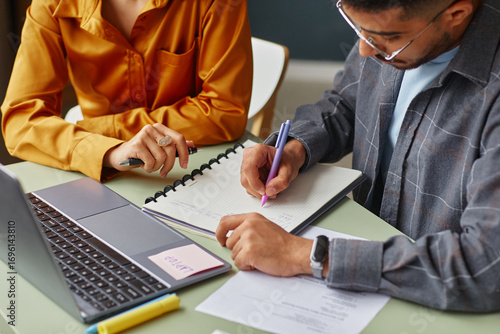 Young adult Caucasian woman and young adult South Asian man studying together at table, writing in notebook, using laptop and reviewing documents in language school classroom setting