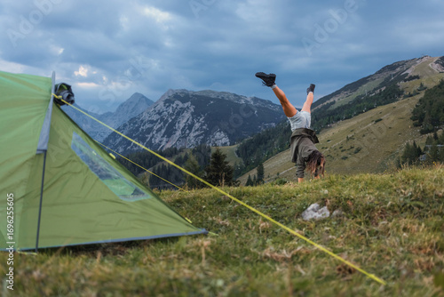Konstfotografi A person performs a handstand in a grassy mountainous area near a tent under a cloudy sky, showcasing the joy of outdoor adventures