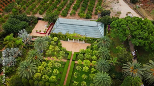Drone video shows a lush public garden in Spain, with structured greenery, a pool, and architectural elements surrounded by orchards and scenic landscapes.