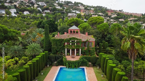 Drone video of a Spanish public garden featuring a historic ivy-covered building, elegant topiary, palm trees, and a pool surrounded by lush greenery.