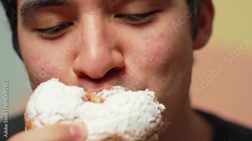 Young Asian man indulgently bites into a fluffy pastry generously covered with powdered sugar, satisfying a sweet craving