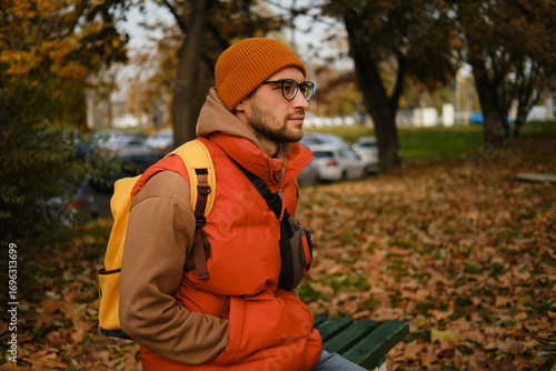 Man with orange beanie and glasses sitting on park bench, hands in pockets and autumn leaves around