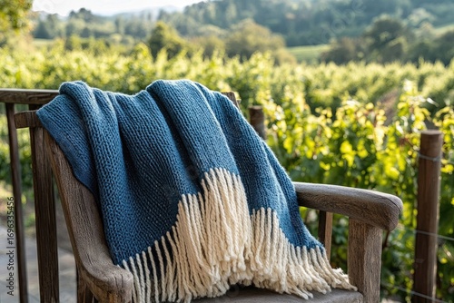 A blue and cream throw blanket with fringes rests on a wooden chair overlooking a vineyard.