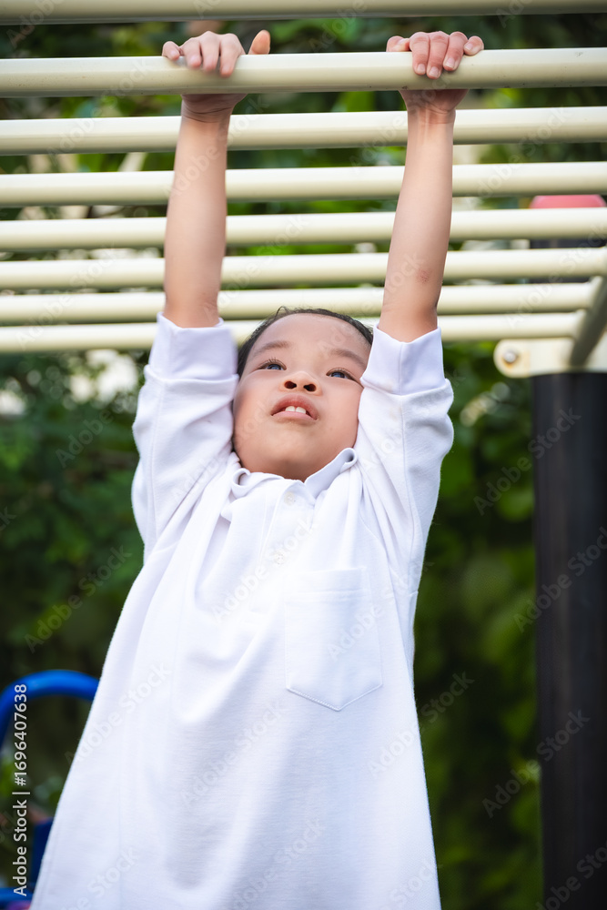Fototapeta premium Portrait of happy young girl hanging on monkey bars at outdoor playground in summer park. Asian girl demonstrating strength and joy while hanging from a bar during sunny day in a vibrant setting.