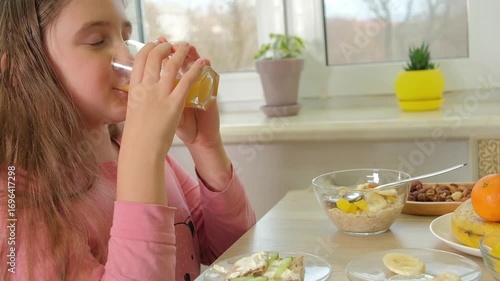 Kid girl in a pink shirt drinks a glass of orange juice. Healthy breakfast and a cheerful morning routine at home
