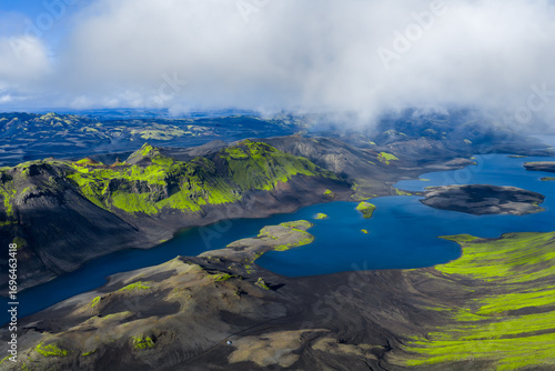 Fotografie Aerial image of Iceland's rugged terrain featuring green moss covered hills, volcanic craters, deep blue lakes, and low hanging clouds